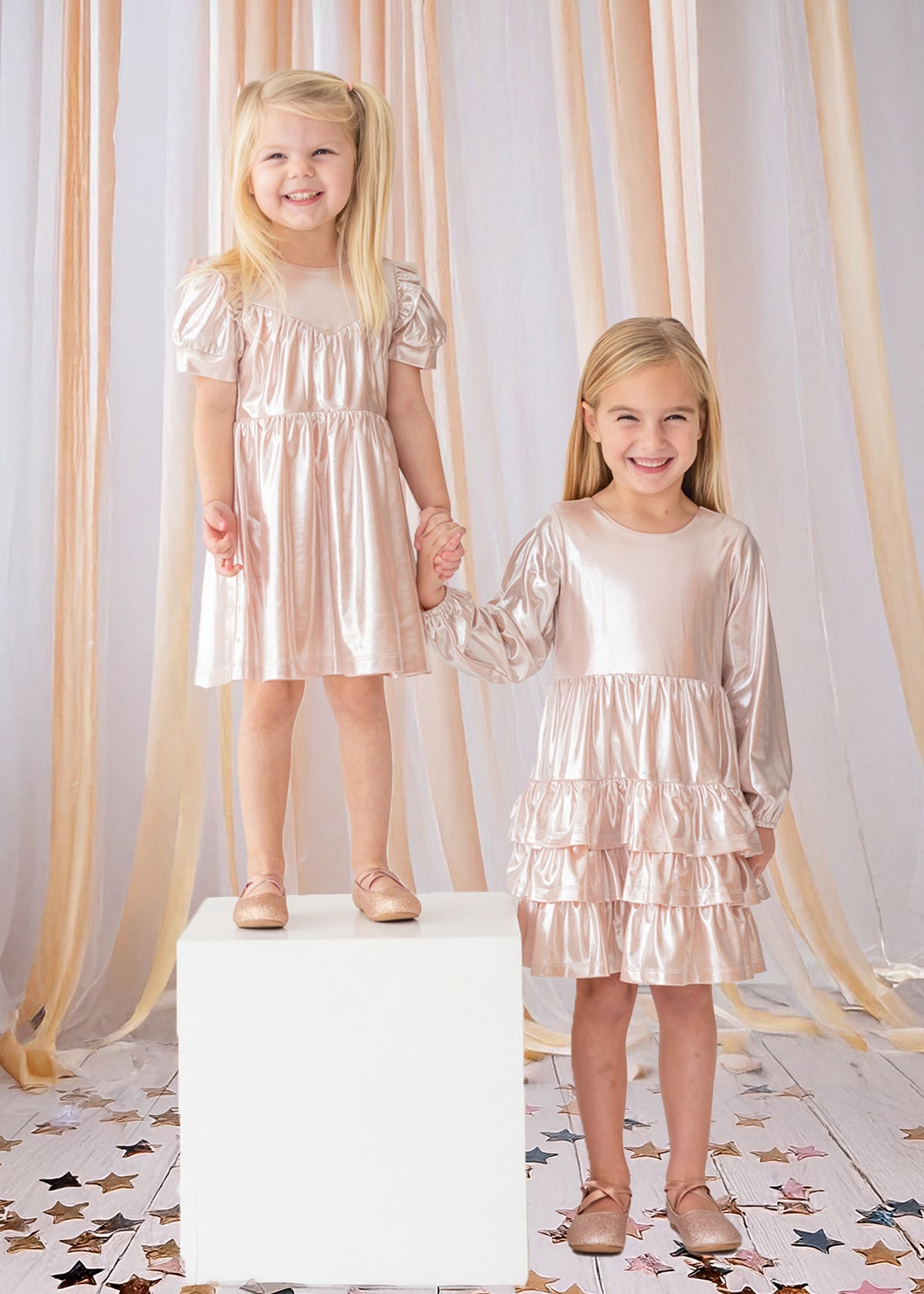 Two young girls hold hands and smile, both wearing Isobella and Chloe’s Golden Glow Pink Dresses with puffed sleeves and matching shoes. One girl stands on a white cube, surrounded by peach ribbons and star confetti for a magical touch.