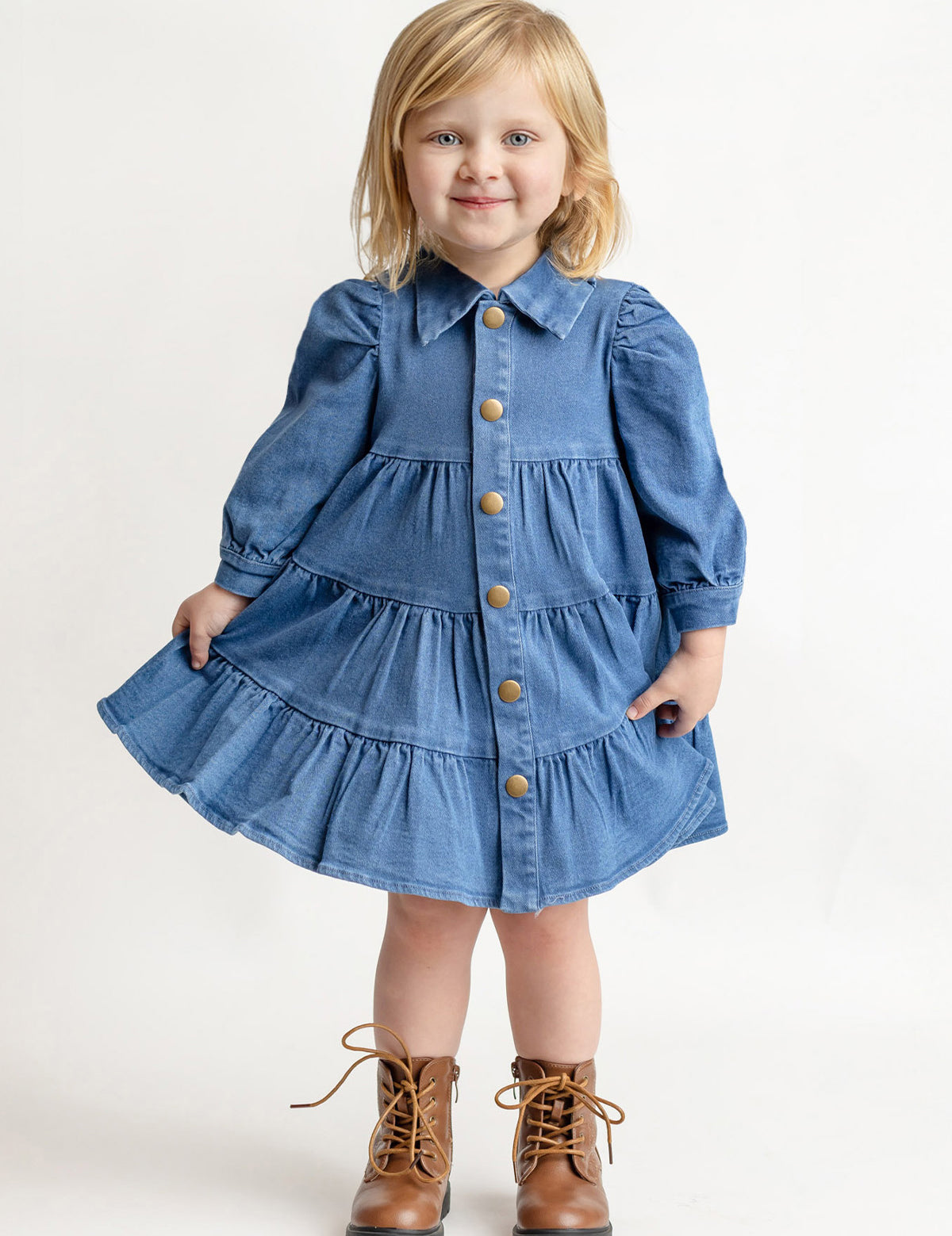 A young blonde-haired child wears the Mabel and Honey Denim Days Tiered Dress—a blue, long-sleeve, button-down denim dress—paired with brown lace-up boots, smiling while standing against a plain white background.