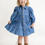 A young blonde-haired child wears the Mabel and Honey Denim Days Tiered Dress—a blue, long-sleeve, button-down denim dress—paired with brown lace-up boots, smiling while standing against a plain white background.