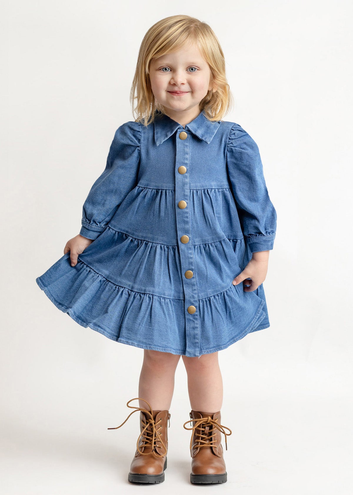 A young blonde-haired child wears the Mabel and Honey Denim Days Tiered Dress—a blue, long-sleeve, button-down denim dress—paired with brown lace-up boots, smiling while standing against a plain white background.