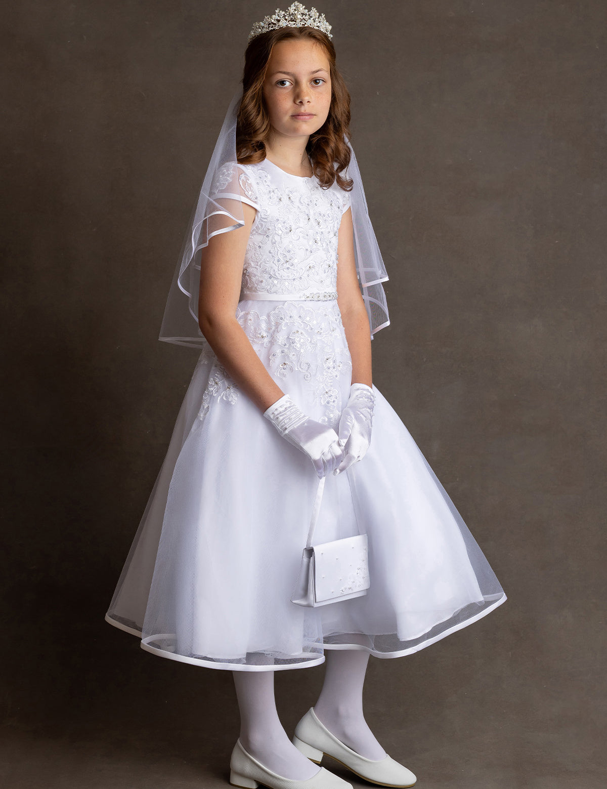 A young girl stands against a plain background, wearing the Lauren Marie Embroidered Bodice and Flower Beaded Waist Dress with gloves, shoes, veil, tiara, and holding a small white purse, looking slightly to the side with a neutral expression.