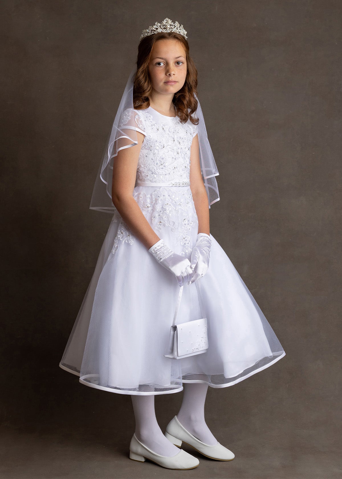 A young girl stands against a plain background, wearing the Lauren Marie Embroidered Bodice and Flower Beaded Waist Dress with gloves, shoes, veil, tiara, and holding a small white purse, looking slightly to the side with a neutral expression.