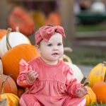 A baby wearing the Mabel and Honey Poppy Rose Long Sleeve Solid Ribbed Bow Tie Bubble Romper in coral, along with a matching headband, sits on grass surrounded by orange and white pumpkins, with additional pumpkins and blurred people visible in the background.