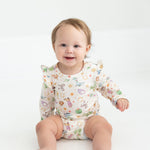 A smiling baby with light brown hair sits on a white floor, wearing the Mabel and Honey Cactus Kisses Long Sleeve Print Ruffle Shoulder Bodysuit.