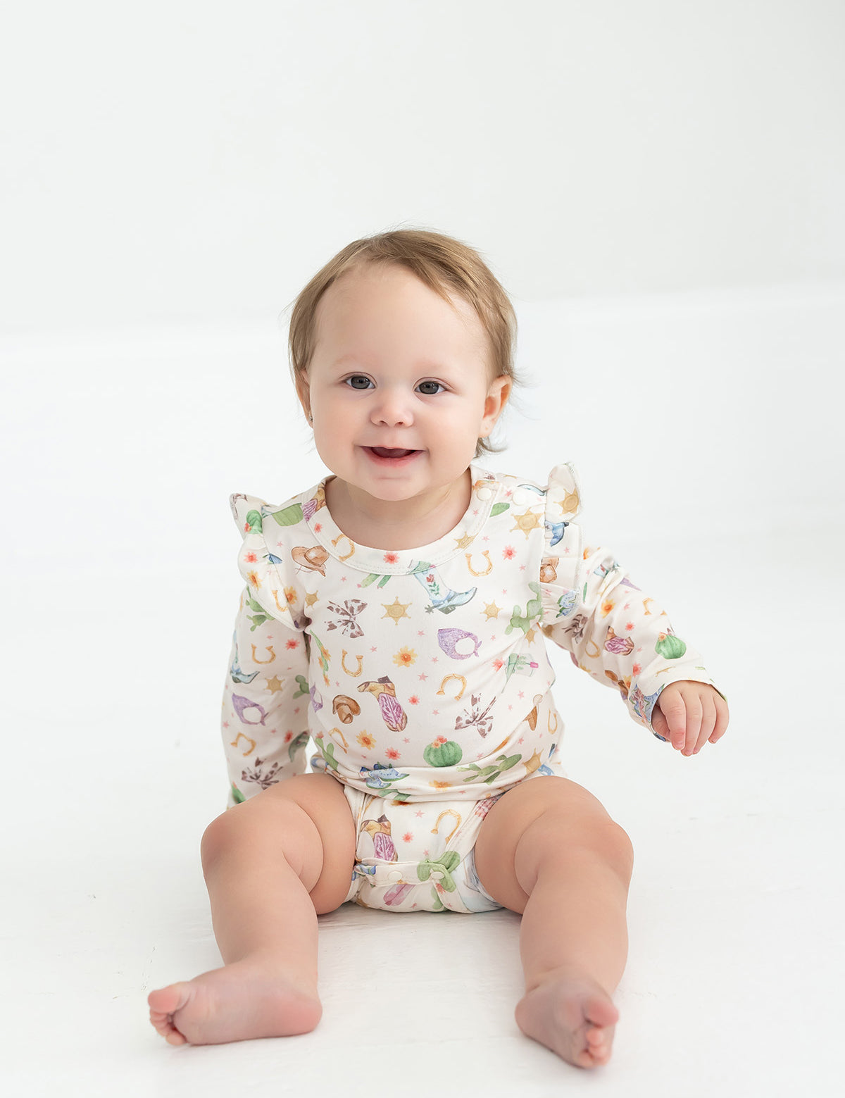 A smiling baby with light brown hair sits on the floor wearing the Mabel and Honey Cactus Kisses Bodysuit Long Sleeve Western Print Ruffle Shoulder Bodysuit, featuring playful western-inspired prints and ruffle shoulder details. The background is white and simple.