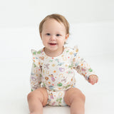 A smiling baby with light brown hair sits on a white floor, wearing the Mabel and Honey Cactus Kisses Long Sleeve Print Ruffle Shoulder Bodysuit.