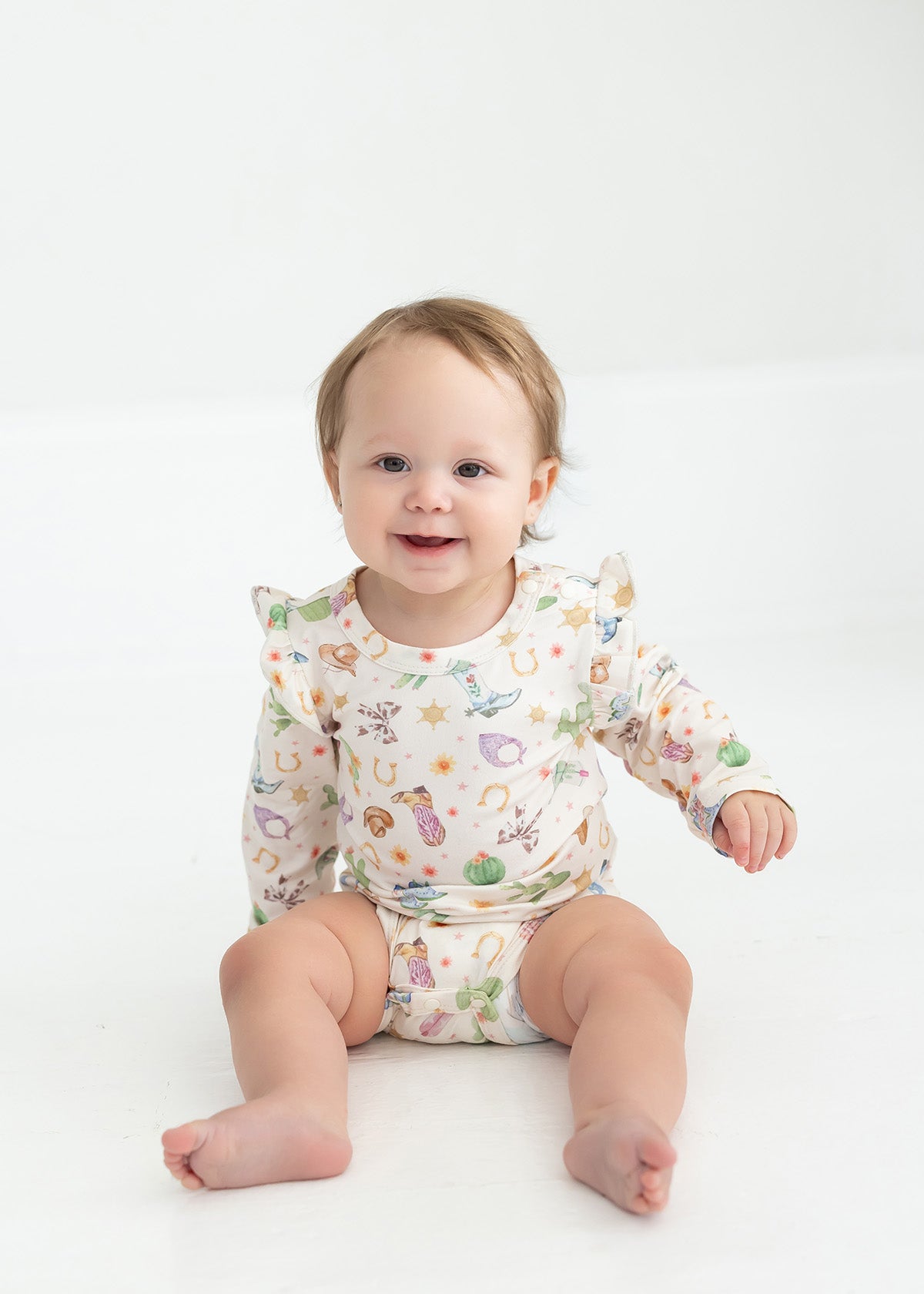A smiling baby with light brown hair sits on a white floor, wearing the Mabel and Honey Cactus Kisses Long Sleeve Print Ruffle Shoulder Bodysuit.