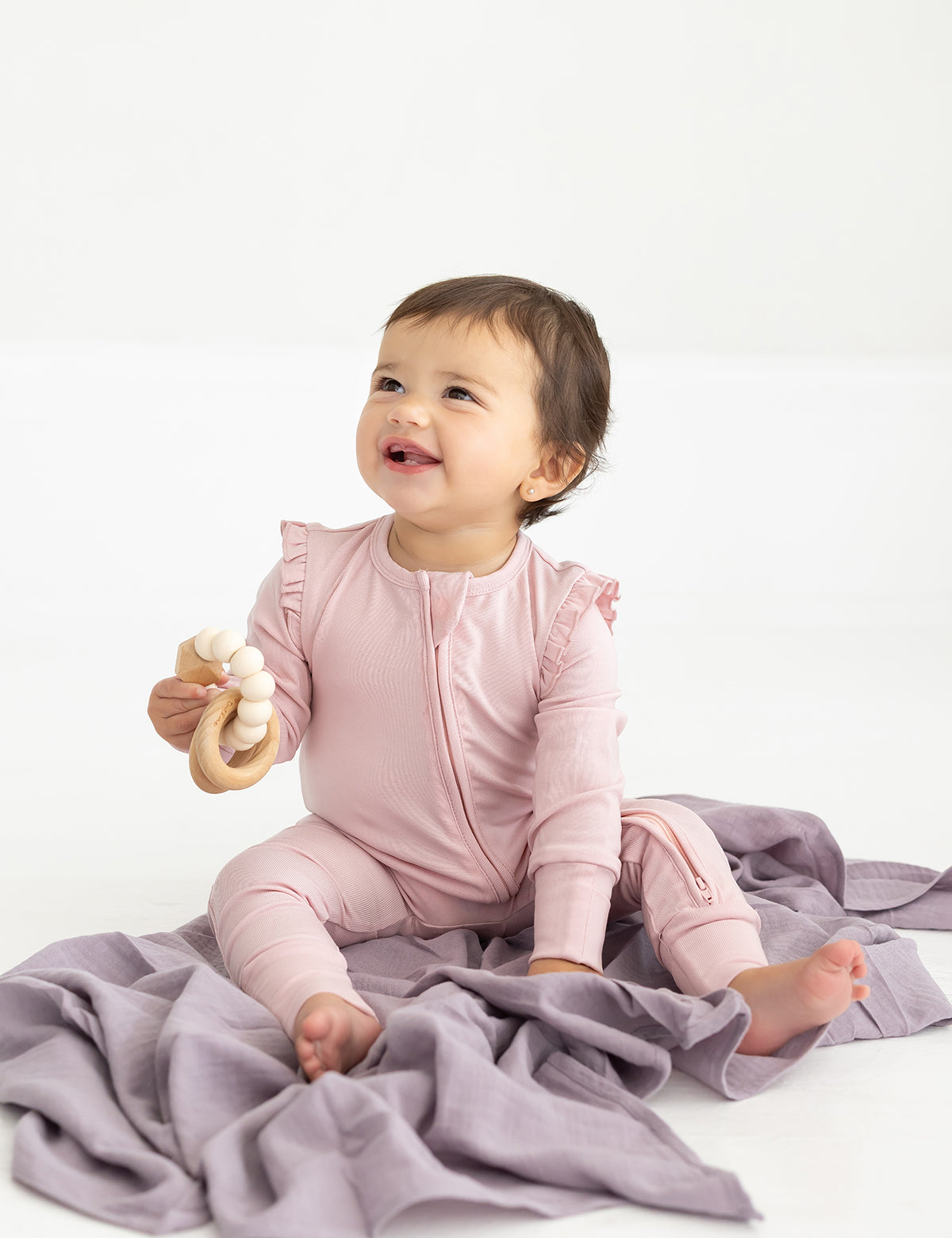 A smiling baby sits on a soft lavender blanket, wearing the Mabel and Honey Whimsical Wildflower Long Sleeve Ruffle Trim Zip Front Convertible Romper and holding a wooden teething toy.