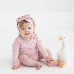 A baby wearing the Mabel and Honey Whimsical Wildflower Long Sleeve Solid Scalloped Trim Bodysuit and a headband with a large bow sits on the floor next to a white and yellow stuffed duck, smiling at the camera.