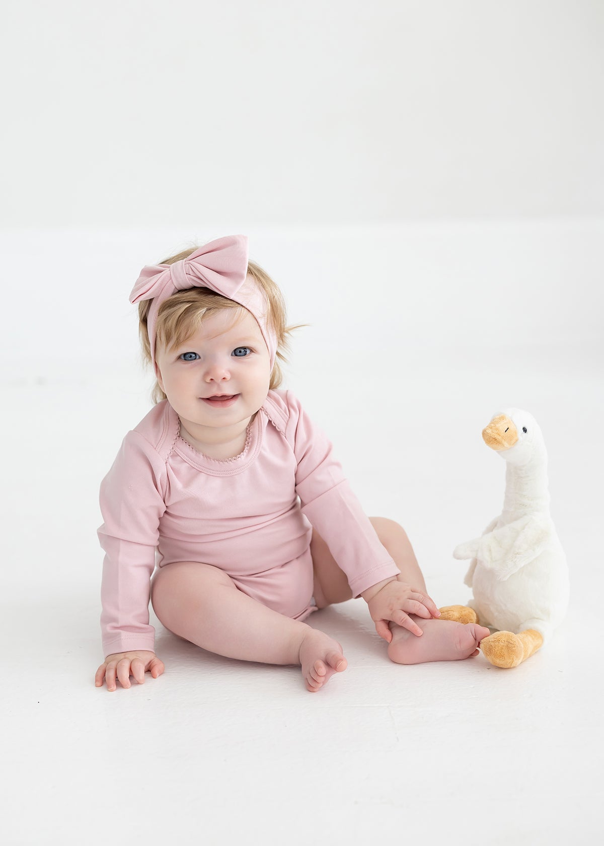 A baby wearing the Mabel and Honey Whimsical Wildflower Long Sleeve Solid Scalloped Trim Bodysuit and a headband with a large bow sits on the floor next to a white and yellow stuffed duck, smiling at the camera.