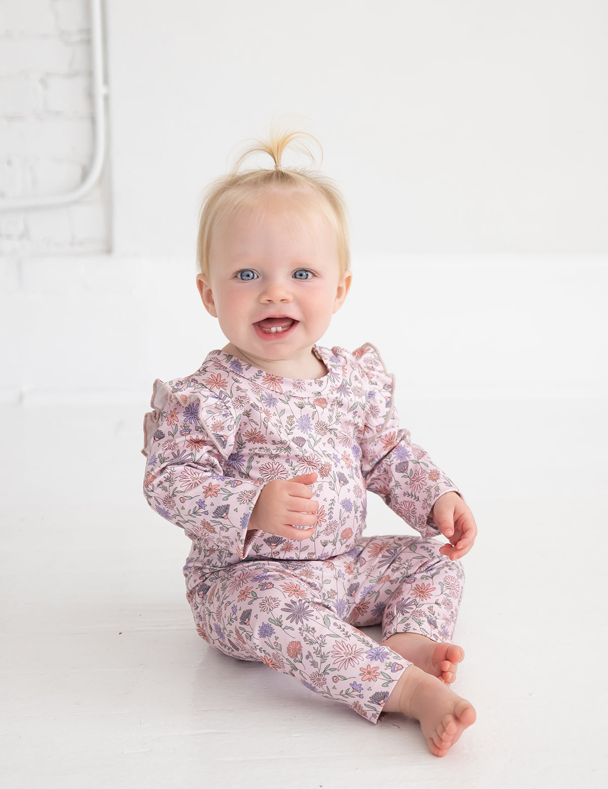 A smiling baby with blond hair in a small ponytail sits barefoot on a white floor against a white wall, wearing the Mabel and Honey Whimsical Wildflower Bodysuit—a long sleeve ruffle shoulder bodysuit adorned with a pink floral print.