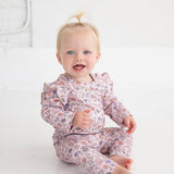 A smiling blonde baby with a top knot sits on the floor in a bright, minimal room with white walls and floors, wearing the Whimsical Wildflower Long Sleeve Floral Ruffle Trim Bodysuit by Mabel and Honey.