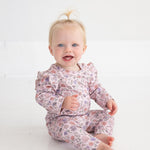 A baby with blonde hair in a small ponytail sits on a white floor, smiling at the camera while wearing Mabel and Honey’s Whimsical Wildflower Floral Elastic Waist Leggings and a matching floral top made from soft stretch knit.