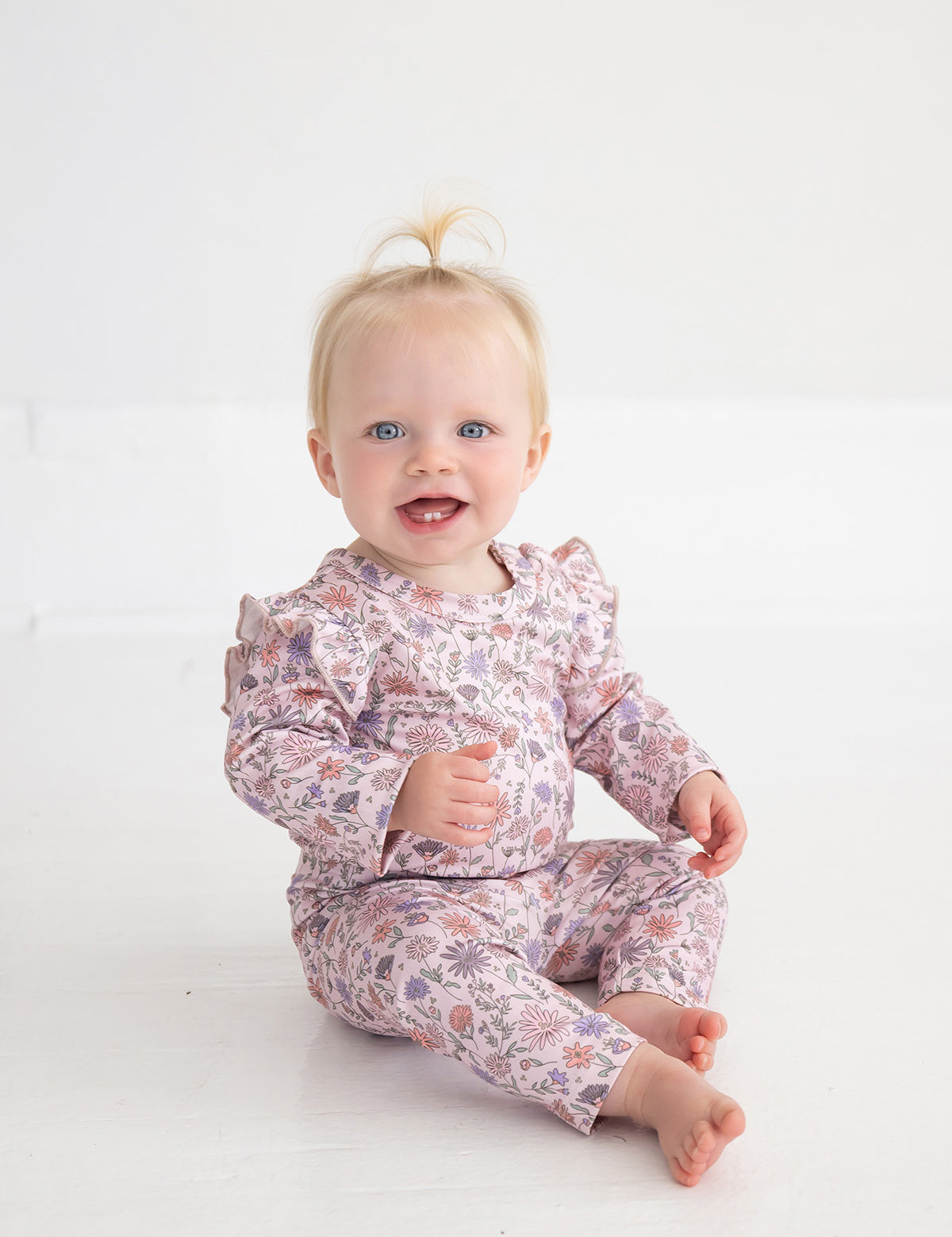A smiling baby with blonde hair sits barefoot on a white floor, wearing Mabel and Honey’s Whimsical Wildflower Legging—straight leg leggings featuring a light pink floral print.
