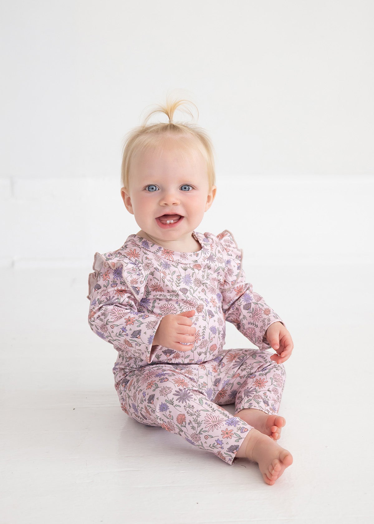 A baby with blonde hair in a small ponytail sits on a white floor, smiling at the camera while wearing Mabel and Honey’s Whimsical Wildflower Floral Elastic Waist Leggings and a matching floral top made from soft stretch knit.