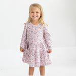 A young girl with blonde hair smiles, standing against a white background. She wears the Mabel and Honey Whimsical Wildflower Long Sleeve Floral Tiered Dress, paired with white socks and matching white shoes.