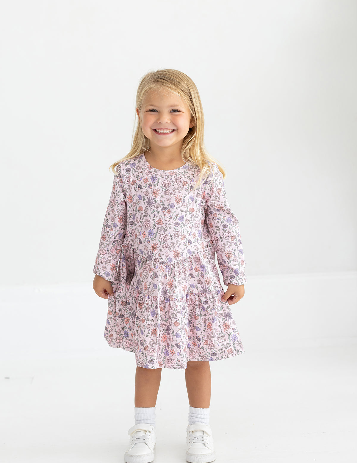 A young girl with long blonde hair smiles while wearing the Mabel and Honey Whimsical Wildflower Dress—a blush pink, long-sleeve, floral tiered A-line dress—paired with white socks and white shoes, standing against a plain white background.