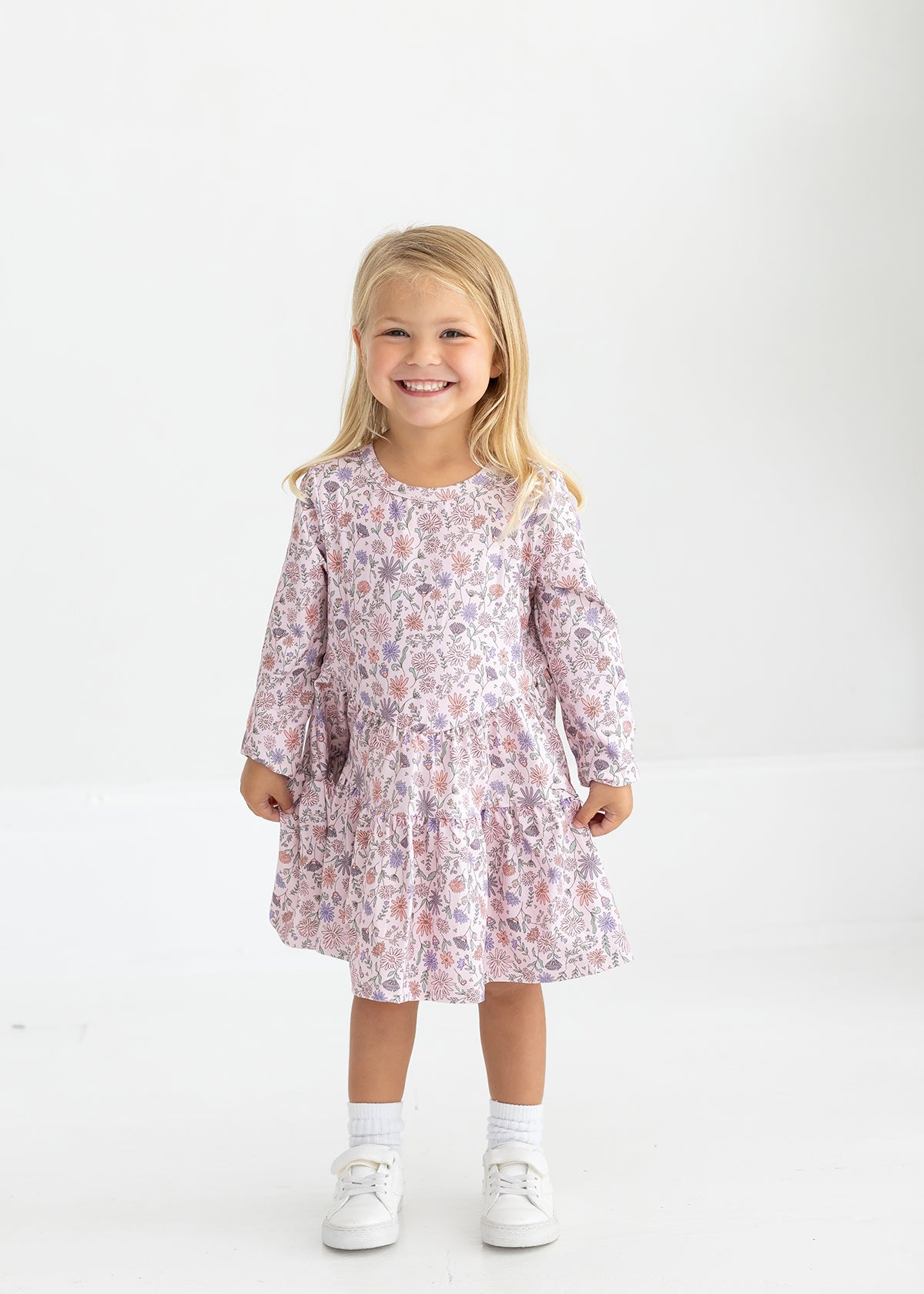 A young girl with blonde hair smiles, standing against a white background. She wears the Mabel and Honey Whimsical Wildflower Long Sleeve Floral Tiered Dress, paired with white socks and matching white shoes.