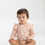 A toddler with short brown hair sits on the floor wearing the Mabel and Honey Honey Petals Long Sleeve Floral Ruffle Shoulder Bodysuit, holding a wooden toy. The plain white background highlights this charming piece of baby girl clothing.