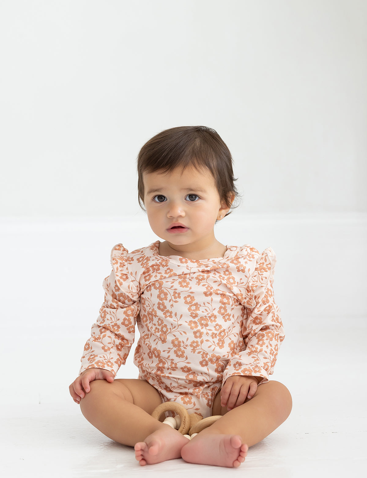 A barefoot toddler with short brown hair sits on a white floor, wearing the Mabel and Honey Honey Petals Bodysuit Long Sleeve Floral Print Ruffle Shoulder Bodysuit. The child gazes forward calmly while holding a small wooden toy.