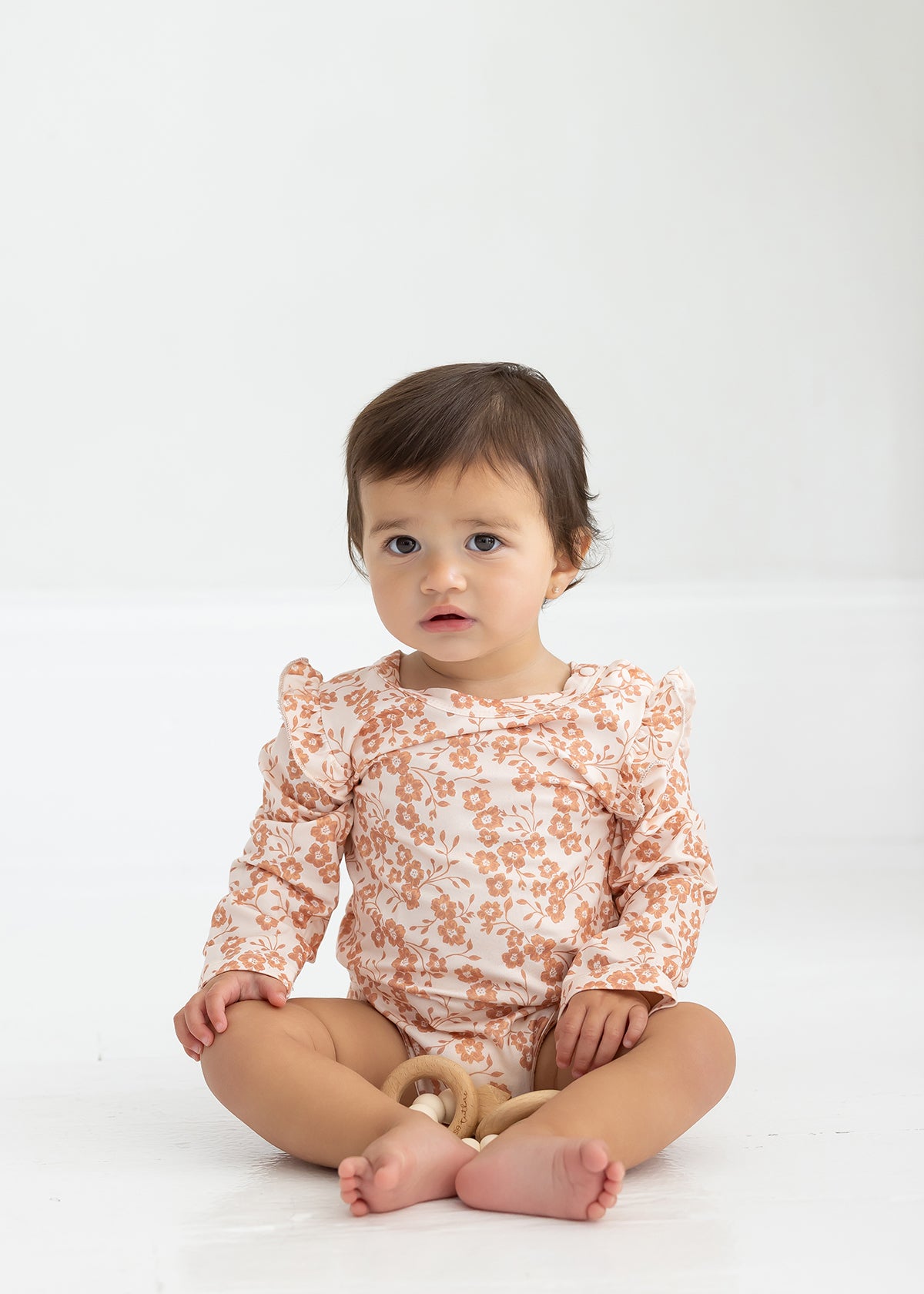 A toddler with short brown hair sits on the floor wearing the Mabel and Honey Honey Petals Long Sleeve Floral Ruffle Shoulder Bodysuit, holding a wooden toy. The plain white background highlights this charming piece of baby girl clothing.