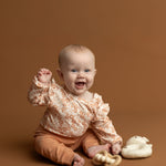 A happy baby wearing the Mabel and Honey Honey Petals Long Sleeve Floral Ruffle Shoulder Bodysuit sits on a brown background, holding a wooden toy, with a soft bunny toy beside them.