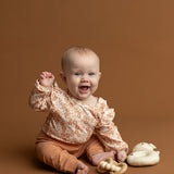 A happy baby wearing the Mabel and Honey Honey Petals Long Sleeve Floral Ruffle Shoulder Bodysuit sits on a brown background, holding a wooden toy, with a soft bunny toy beside them.