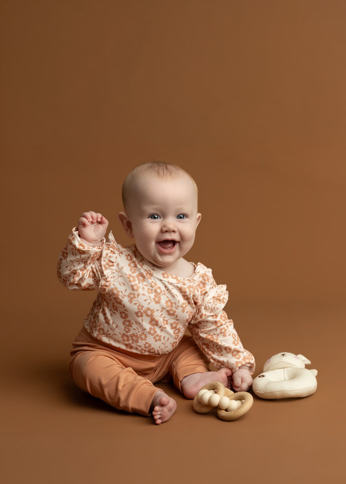 A happy baby wearing the Mabel and Honey Honey Petals Long Sleeve Floral Ruffle Shoulder Bodysuit sits on a brown background, holding a wooden toy, with a soft bunny toy beside them.