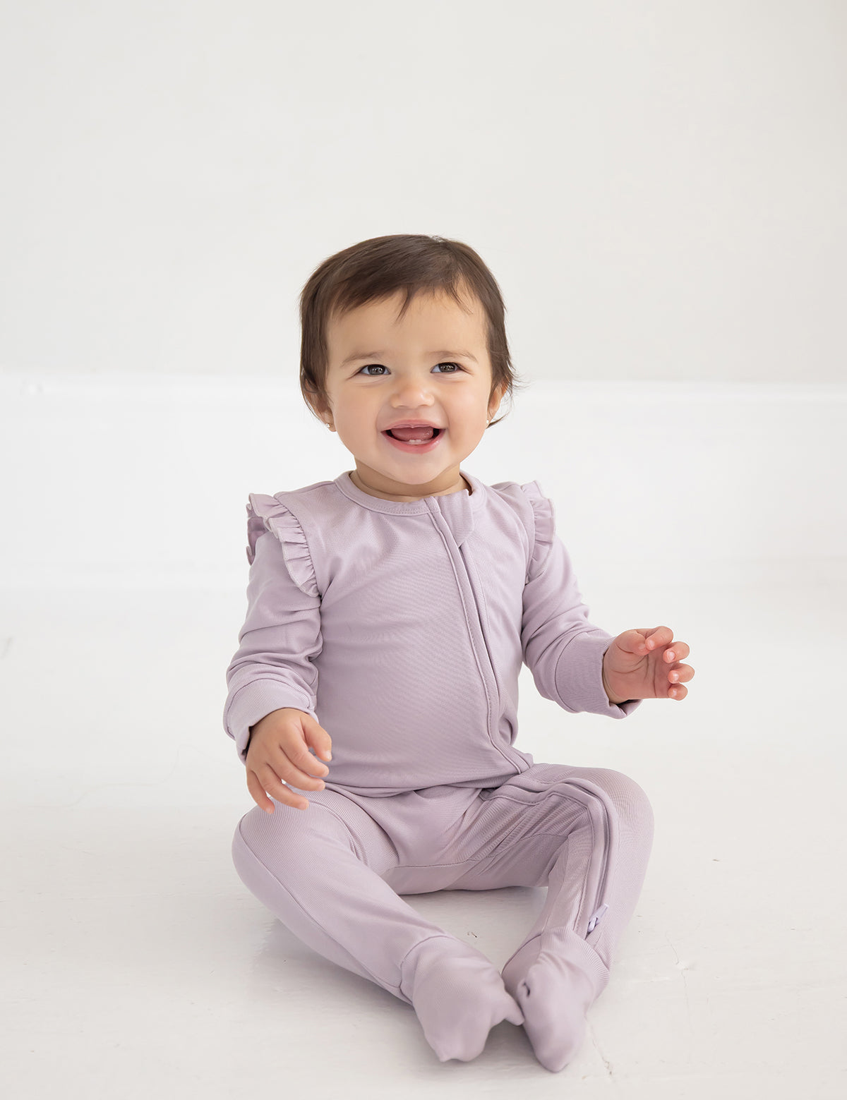 A smiling toddler with short brown hair sits on the floor, wearing the Mabel and Honey Soft Meadow Convertible Romper— a long sleeve, solid straight leg romper crafted from soft Tencel knit—against a plain white background.