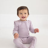 A smiling baby with short brown hair sits on a white floor, wearing the Soft Meadow Ruffle Trim Convertible Foot Long Sleeve Solid Romper by Mabel and Honey—a light purple, long-sleeve romper crafted from soft ribbed stretch fabric and finished with ruffled shoulders.