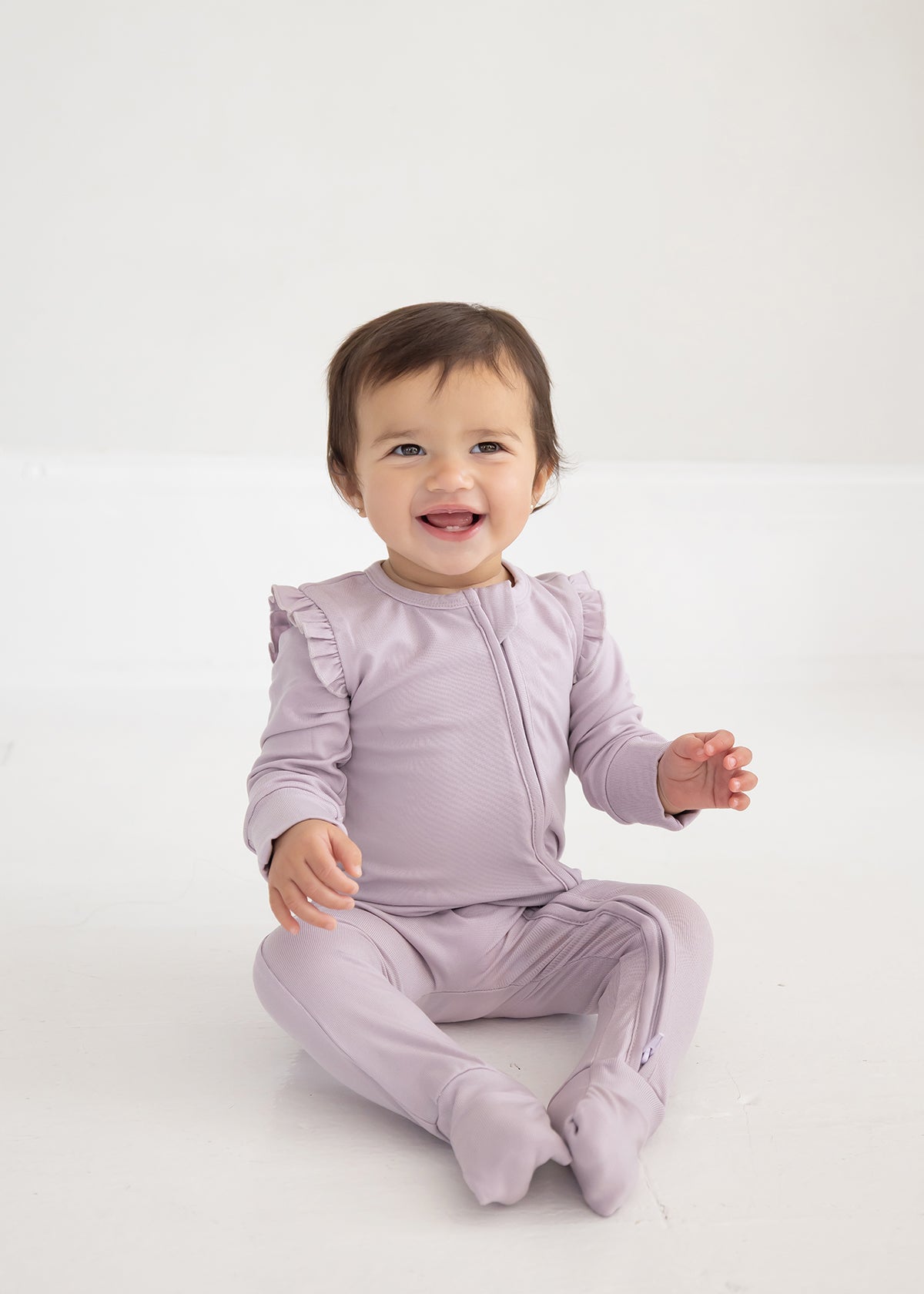 A smiling baby with short brown hair sits on a white floor, wearing the Soft Meadow Ruffle Trim Convertible Foot Long Sleeve Solid Romper by Mabel and Honey—a light purple, long-sleeve romper crafted from soft ribbed stretch fabric and finished with ruffled shoulders.