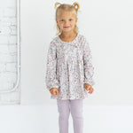 A young child with blonde hair in pigtails is standing and smiling against a white background, wearing the Mabel and Honey Soft Meadow Long Sleeve Floral Peter Pan Collar Top and Legging Set, paired with white sneakers.