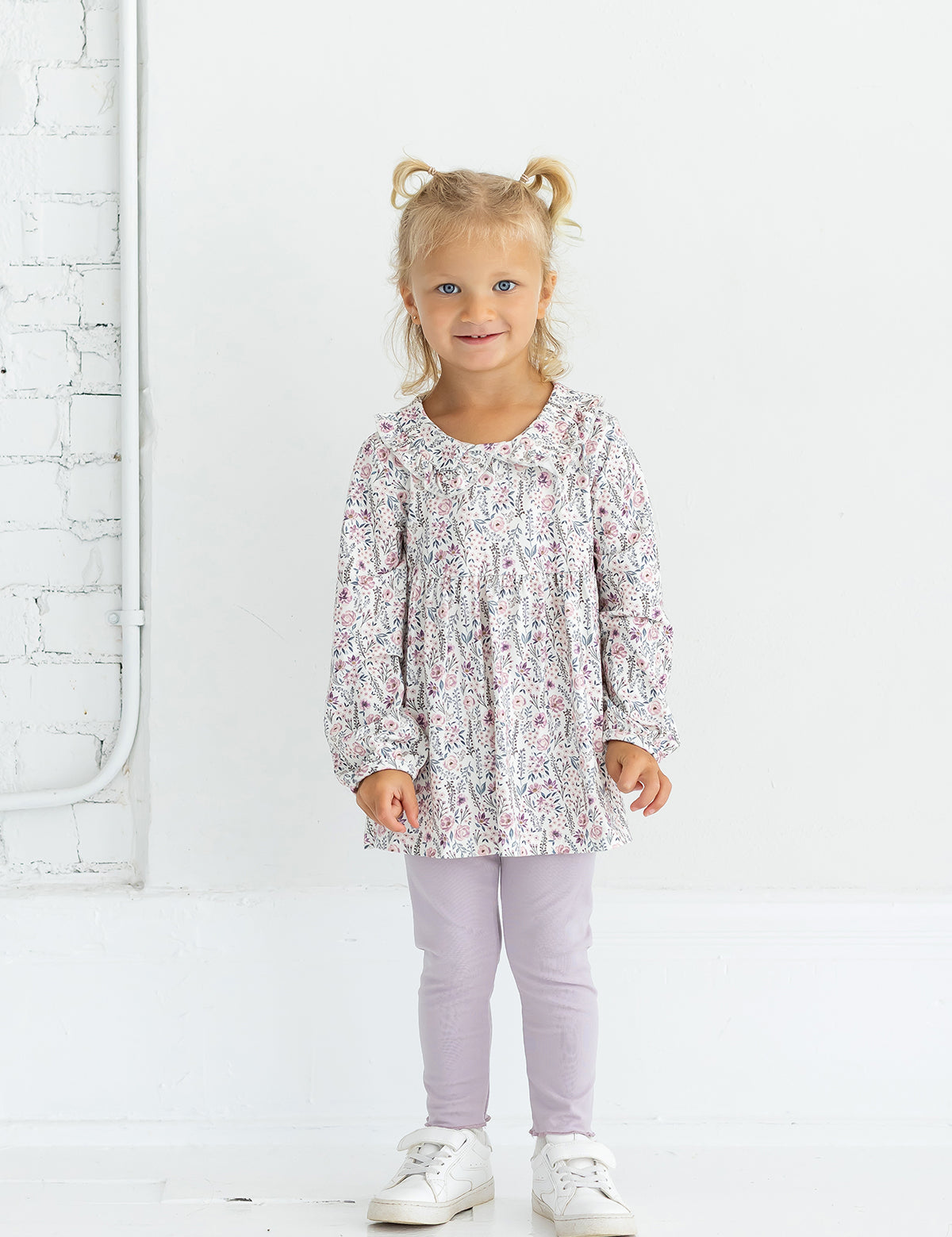 A young child with blonde hair in pigtails is standing and smiling against a white background, wearing the Mabel and Honey Soft Meadow Long Sleeve Floral Peter Pan Collar Top and Legging Set, paired with white sneakers.