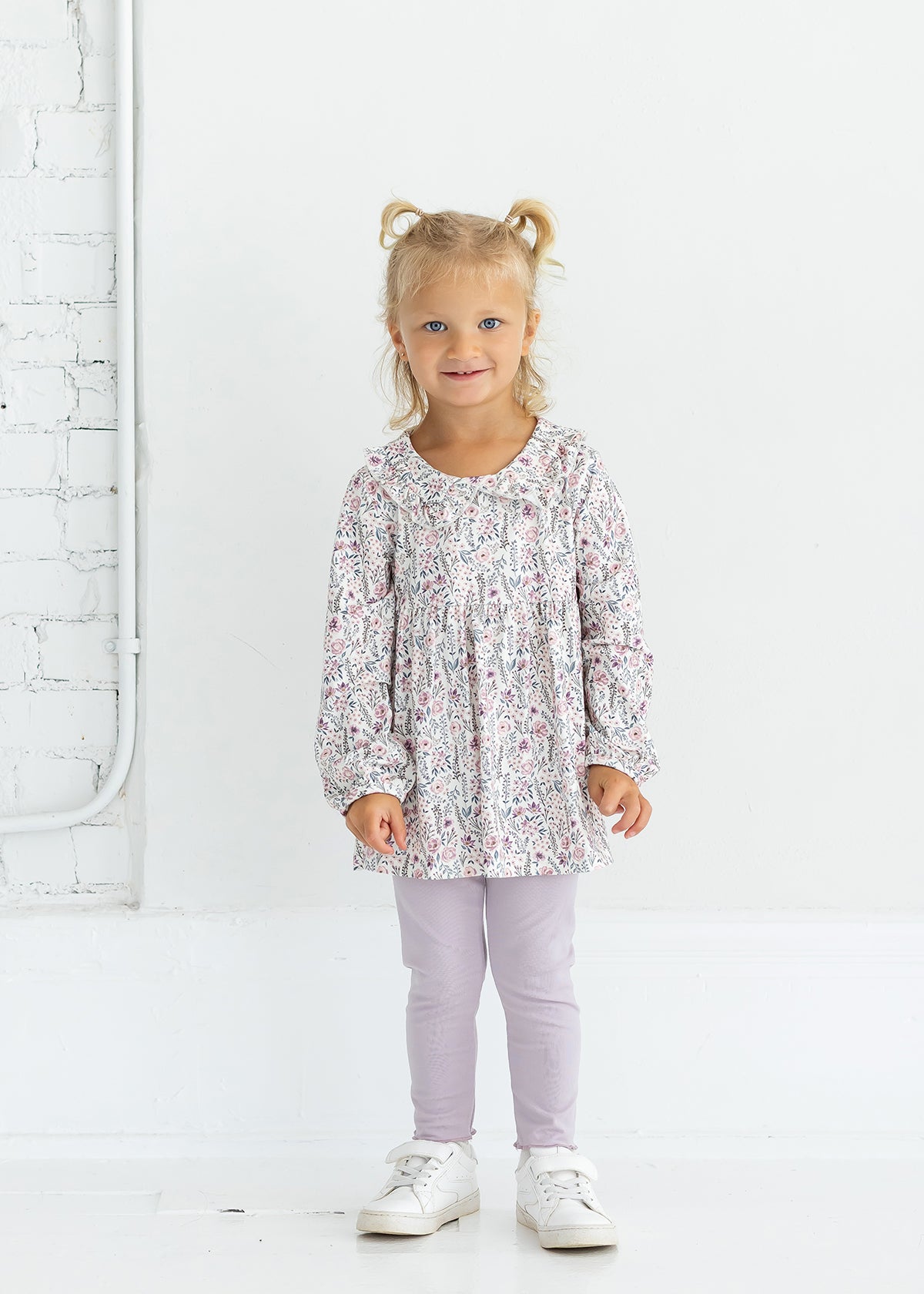 A young child with blonde hair in pigtails is standing and smiling against a white background, wearing the Mabel and Honey Soft Meadow Long Sleeve Floral Peter Pan Collar Top and Legging Set, paired with white sneakers.
