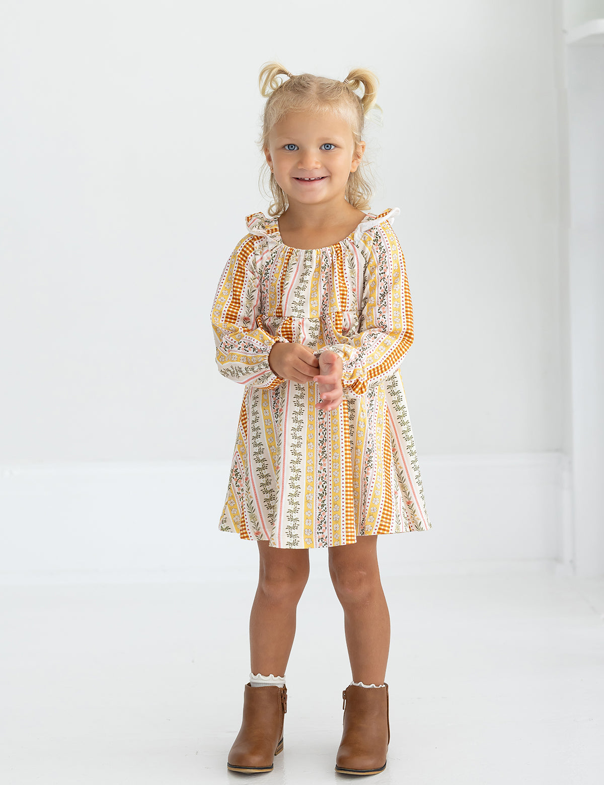 A young child with blonde hair in pigtails smiles and stands in a white room, wearing the Mabel and Honey Buttercup Blossom Long Sleeve Print Ruffle Tie Shoulder A Line Dress paired with brown ankle boots.