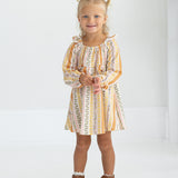 A young child with blonde hair in pigtails smiles and stands in a white room, wearing the Mabel and Honey Buttercup Blossom Long Sleeve Print Ruffle Tie Shoulder A Line Dress paired with brown ankle boots.