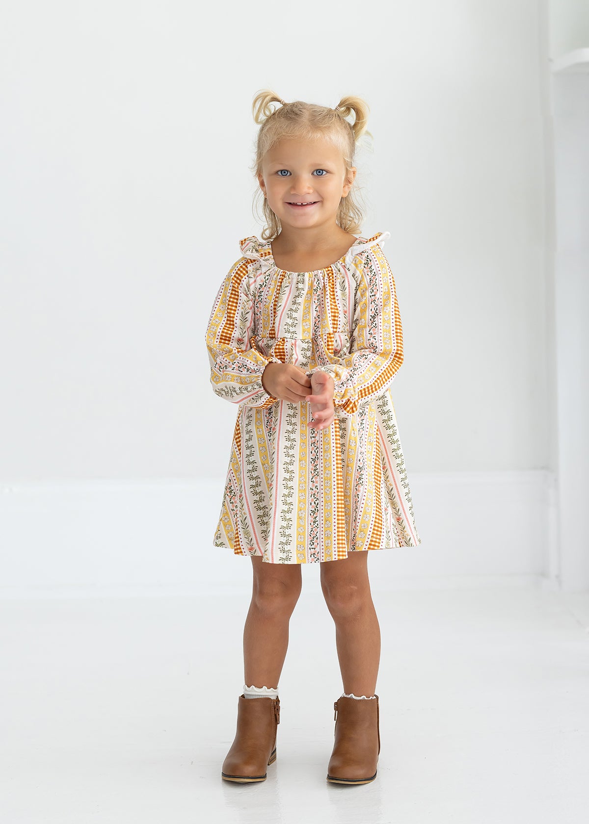 A young child with blonde hair in pigtails smiles and stands in a white room, wearing the Mabel and Honey Buttercup Blossom Long Sleeve Print Ruffle Tie Shoulder A Line Dress paired with brown ankle boots.