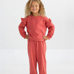 A young child with curly hair stands smiling with hands on hips against a plain white background, wearing the Mabel and Honey Red Rosie Long Sleeve Solid Ruffle Trim Cropped Sweater paired with matching pants.