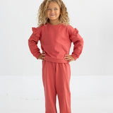 A young child with curly hair stands smiling with hands on hips against a plain white background, wearing the Mabel and Honey Red Rosie Long Sleeve Solid Ruffle Trim Cropped Sweater paired with matching pants.