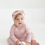 A baby with blonde hair and blue eyes wears a pink headband with a bow and a matching pink outfit, featuring Mabel and Honey’s Whimsical Wildflower Ribbed Waistband Cuffed Joggers in a soft cotton blend, while sitting on the floor holding a cuddly toy.