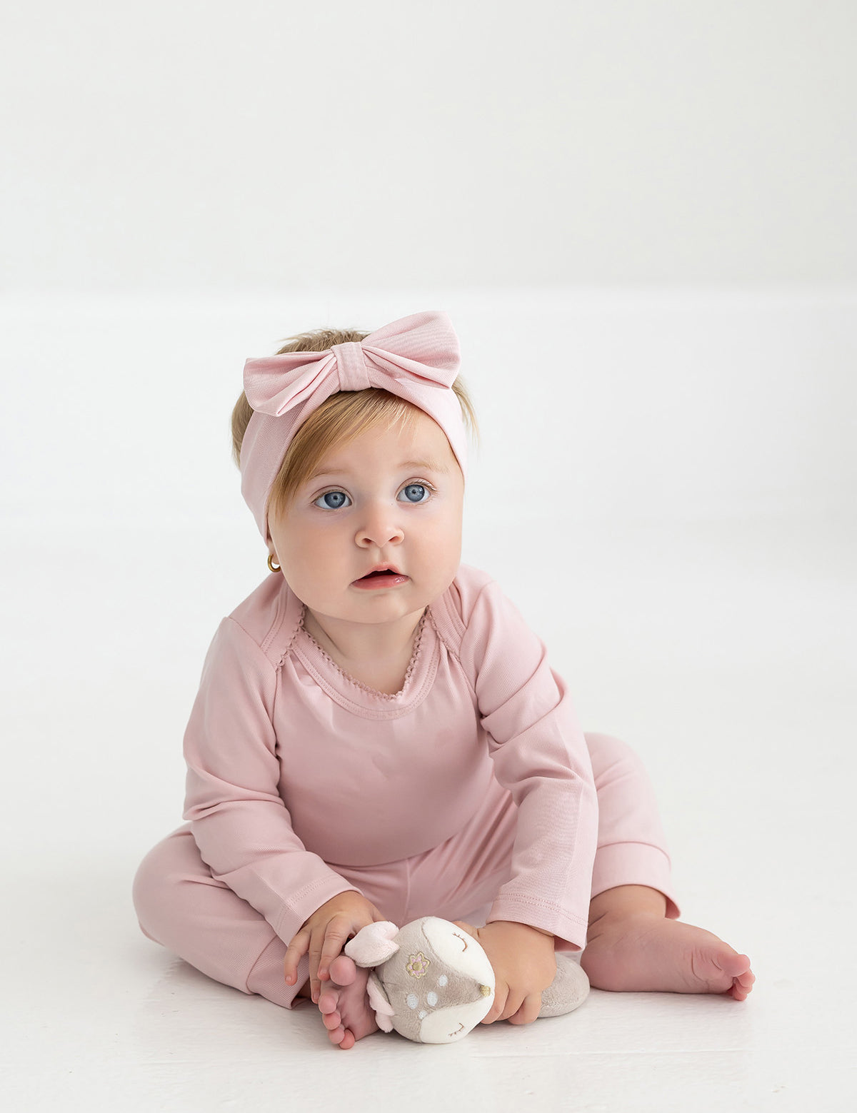 A baby dressed in Mabel and Honey’s Whimsical Wildflower Joggers Ribbed Elastic Waist Cuffed Jogger Leg Joggers and a matching large-bow headband sits on the floor, holding a soft stuffed toy against a plain white background.