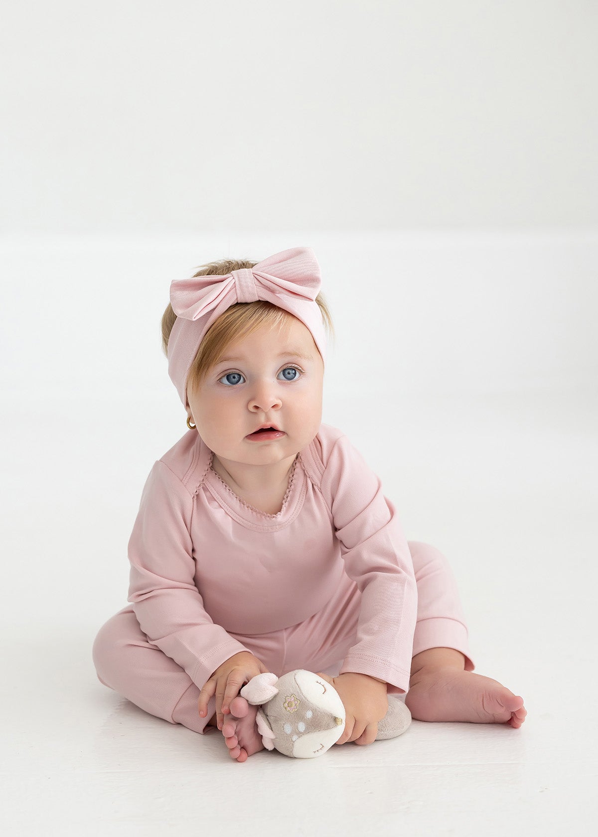 A baby with blonde hair and blue eyes wears a pink headband with a bow and a matching pink outfit, featuring Mabel and Honey’s Whimsical Wildflower Ribbed Waistband Cuffed Joggers in a soft cotton blend, while sitting on the floor holding a cuddly toy.
