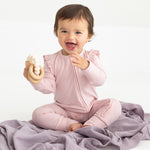 A smiling baby sits on a lavender blanket wearing the Mabel and Honey Whimsical Wildflower Long Sleeve Convertible Romper with Headband in soft pink, holding a cream-colored teething toy.