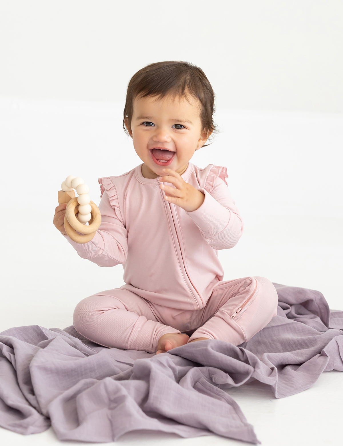 A smiling baby sits on a lavender blanket wearing the Mabel and Honey Whimsical Wildflower Long Sleeve Convertible Romper with Headband in soft pink, holding a cream-colored teething toy.