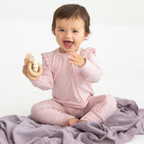 A smiling baby sits on a lavender blanket wearing the Mabel and Honey Whimsical Wildflower Long Sleeve Convertible Romper with Headband in soft pink, holding a cream-colored teething toy.