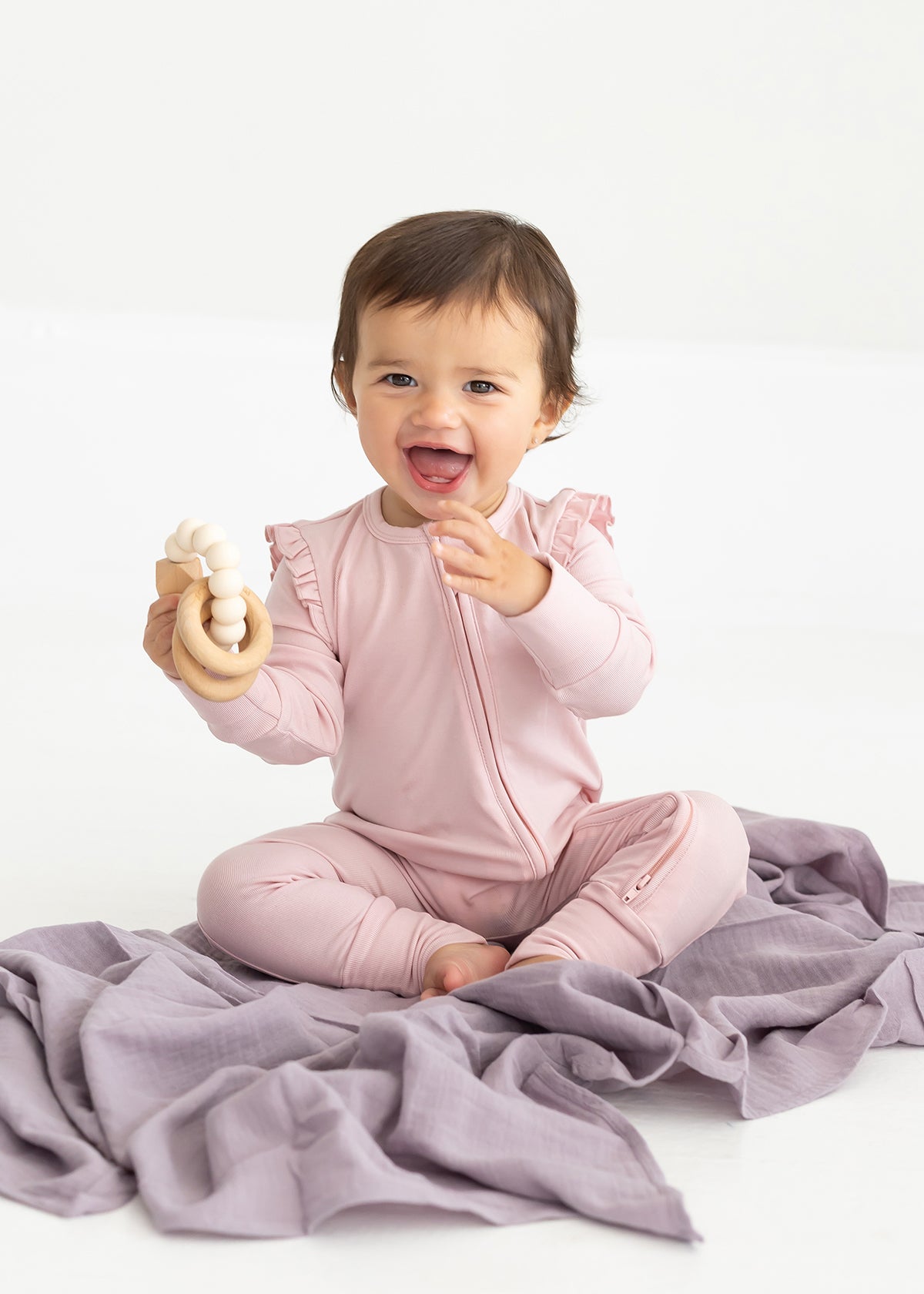 A smiling baby sits on a lavender blanket wearing the Mabel and Honey Whimsical Wildflower Long Sleeve Convertible Romper with Headband in soft pink, holding a cream-colored teething toy.