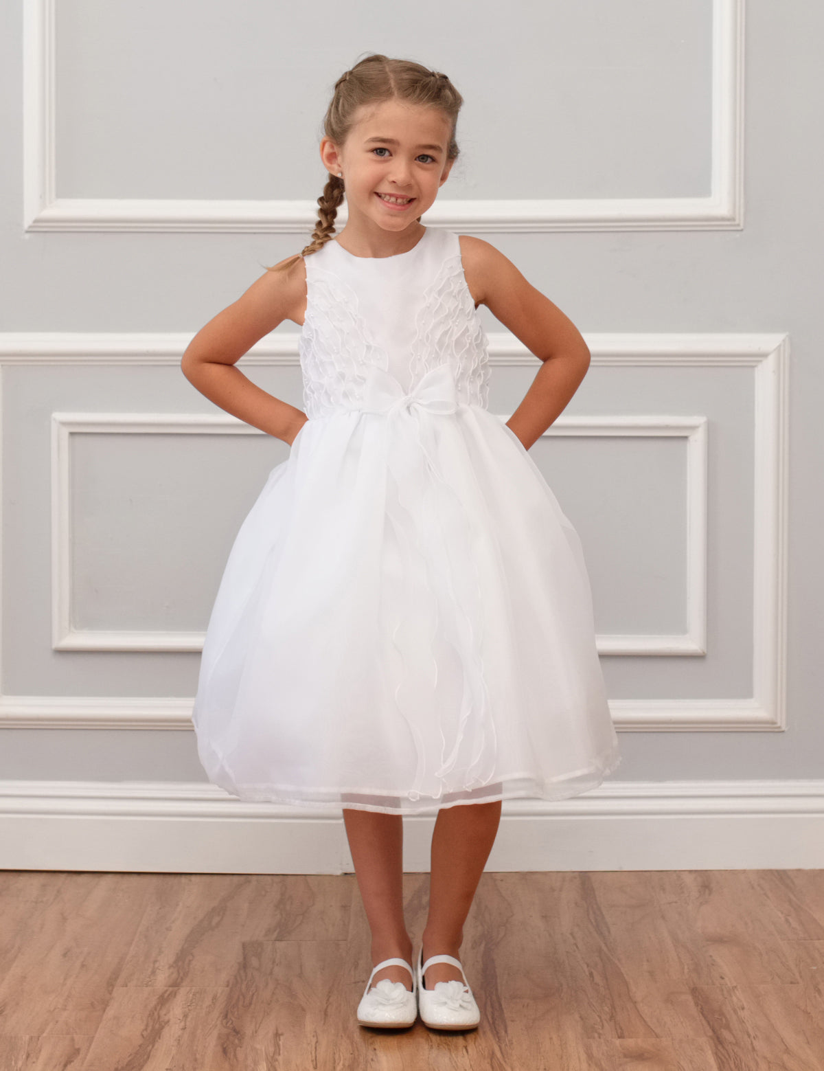A young girl with braided hair smiles against a white paneled wall wearing the Mabel and Honey Sleeveless Organza Communion and Flower Girl Bow Dress, featuring a satin bodice, tulle skirt, and bow at the waist, with white shoes on a wooden floor.