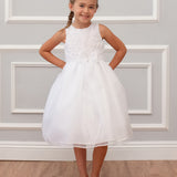 A young girl with braided hair smiles against a white paneled wall wearing the Mabel and Honey Sleeveless Organza Communion and Flower Girl Bow Dress, featuring a satin bodice, tulle skirt, and bow at the waist, with white shoes on a wooden floor.