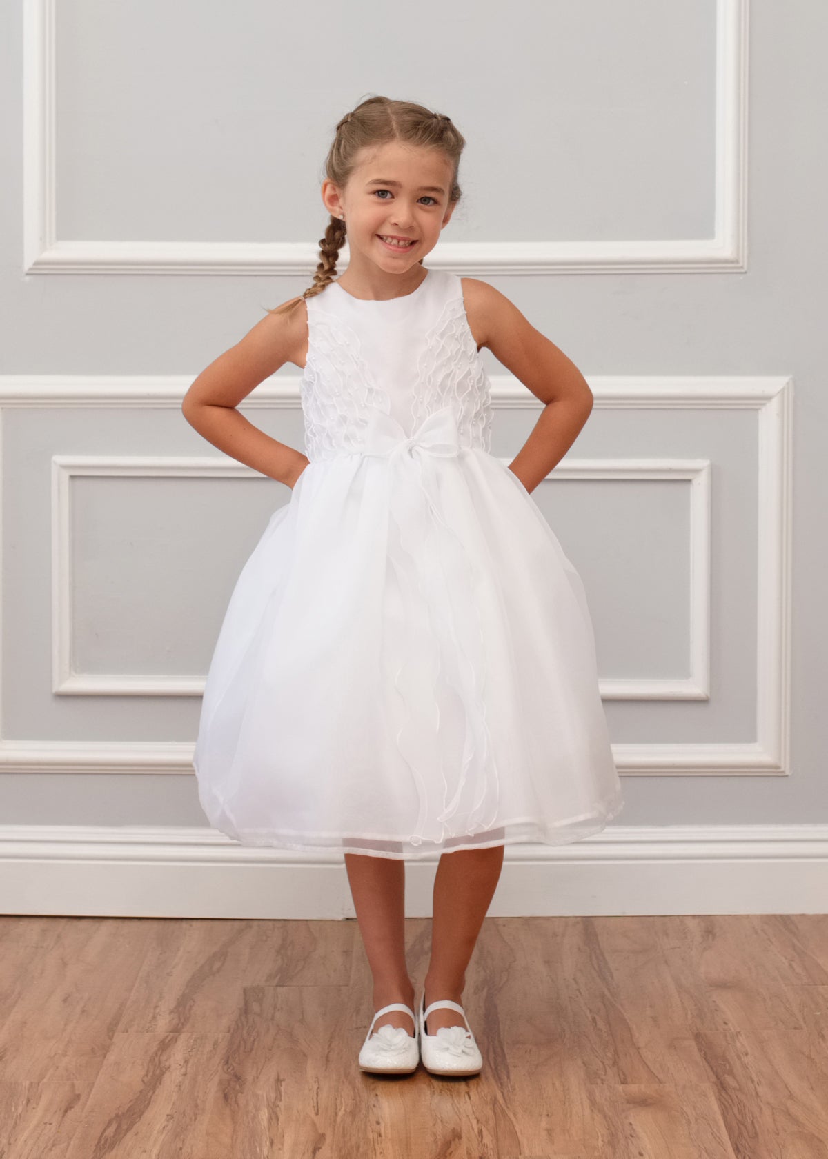 A young girl with braided hair smiles against a white paneled wall wearing the Mabel and Honey Sleeveless Organza Communion and Flower Girl Bow Dress, featuring a satin bodice, tulle skirt, and bow at the waist, with white shoes on a wooden floor.