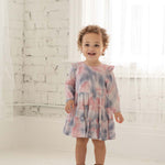 A smiling toddler with curly hair wears the Mabel + Honey Purple Haze Rayon Dress, featuring long sleeves and a tiered tie-dye design, standing in a bright white room with sheer curtains and a painted brick wall.